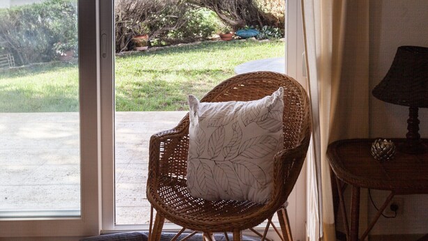 Une chaise en osier avec un coussin, une petite table avec une lampe, et une vue sur la mer par une porte-fenêtre coulissante.