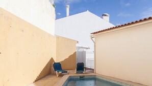 Un petit espace piscine avec deux chaises longues, un sol carrelé et un bâtiment blanc surmonté d’un toit rouge.
