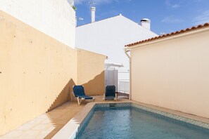 Ein kleiner Pool im Freien mit zwei Liegestühlen, ein weißes Gebäude und ein strahlend blauer Himmel.