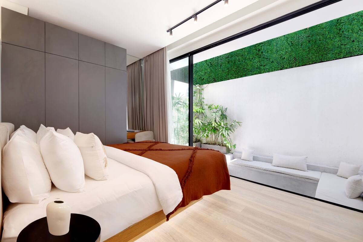 A modern bedroom with a large bed, a bedside table, a potted plant, and a view of greenery through a sliding glass door.