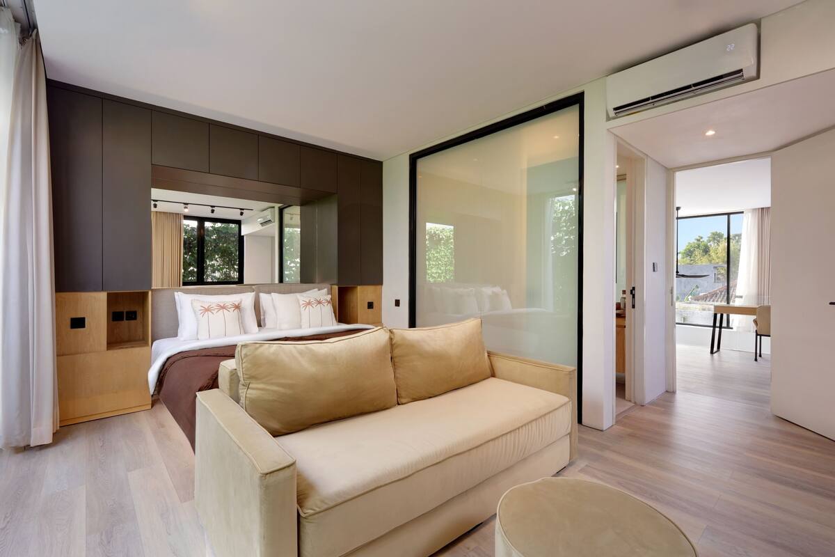 A modern hotel room with a large bed, a sofa, and a dining area visible through a glass wall.