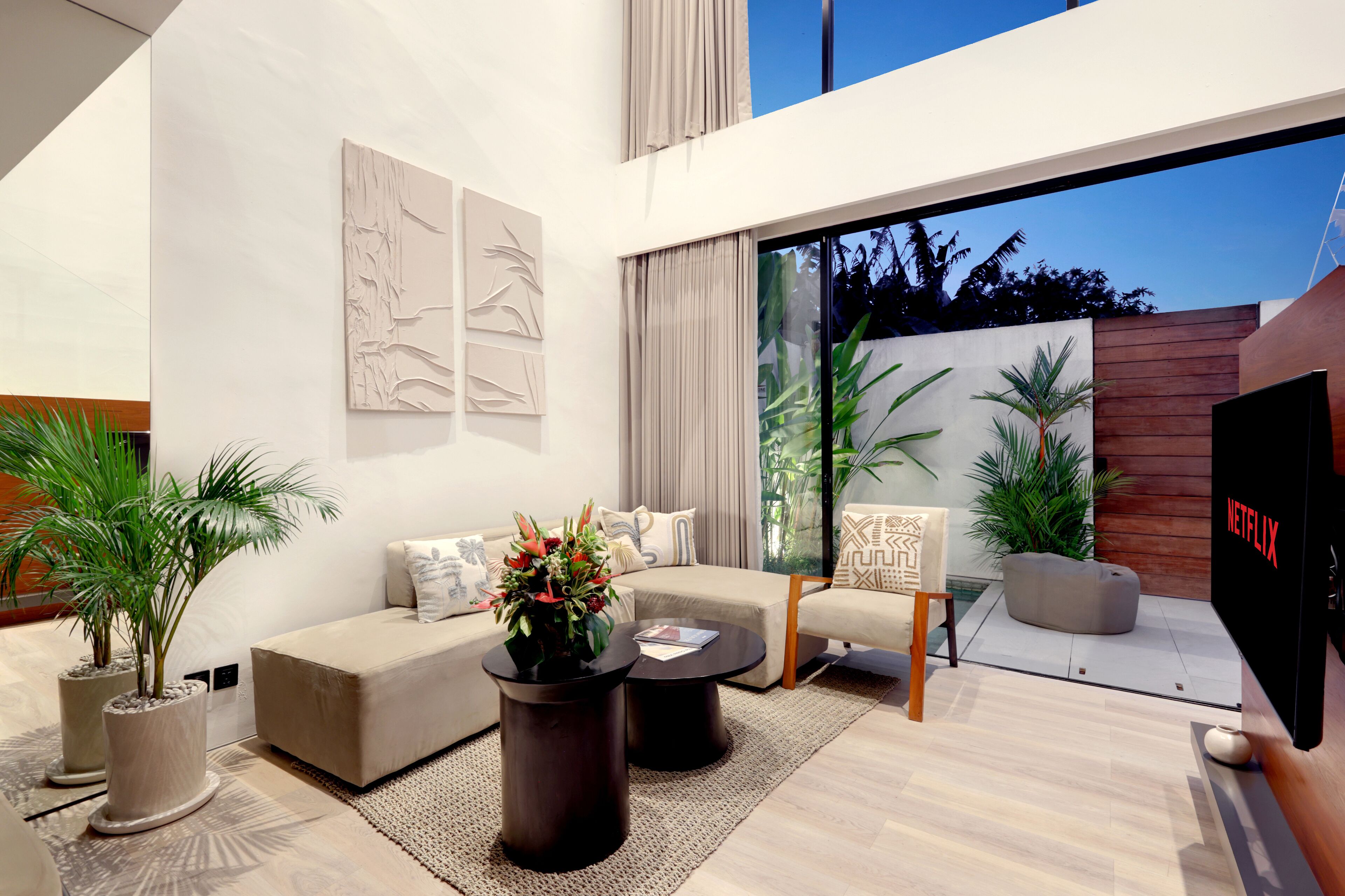 A modern living room with a sofa, coffee table, and chairs, a flat-screen TV, and large windows with a view of greenery.
