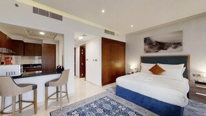 Ein modernes Hotelzimmer mit Küchenzeile, einem großen Bett mit blauem Akzent, einem Bild an der Wand und einem gemusterten Teppich.
