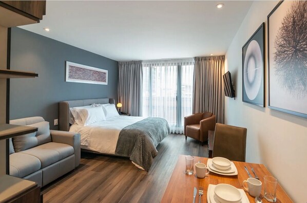 Relax in a stylish space with a neatly made bed, sofa, and dining table, surrounded by artwork and natural light.