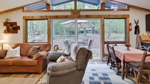 A cozy cabin interior with a wooden ceiling, a leather sofa, a dining table with chairs, and large windows overlooking a forested area. - KingBed+Cabin+quiet+new renovation+foliage+ski (Lake Harmony)