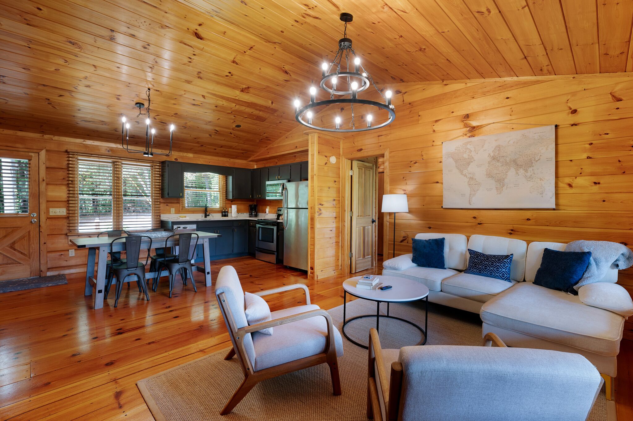 L’intérieur d’un chalet accueillant, avec des murs en bois, un coin repas avec des chaises, un ensemble de canapés et une cuisine visible à travers une porte ouverte.