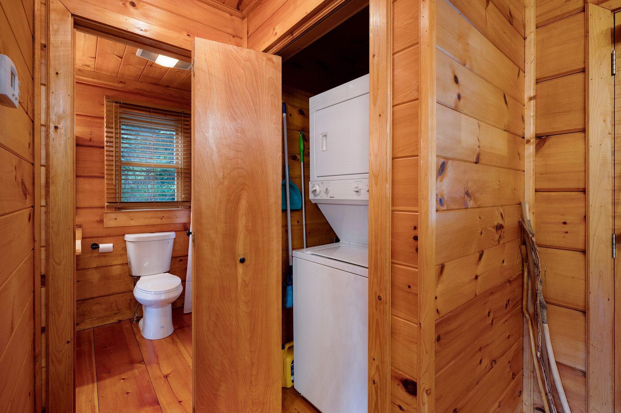 A wooden cabin bathroom with a toilet, washing machine, and a window with blinds.
