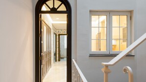 Un pasillo con una escalera que conduce a un segundo piso, una ventana con vista al interior y una puerta al final del pasillo.