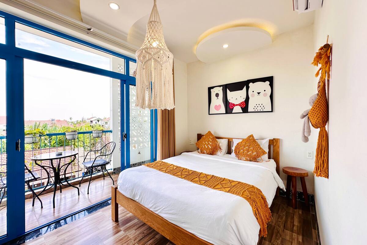 A bedroom with a bed, a hanging macrame light fixture, a wall art piece, and a balcony with a table and chairs.