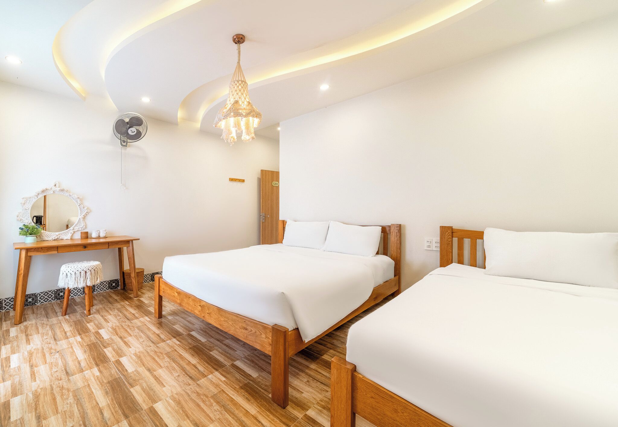 Une chambre d’hôtel avec deux lits, un bureau en bois, une chaise, un miroir et un ventilateur de plafond.
