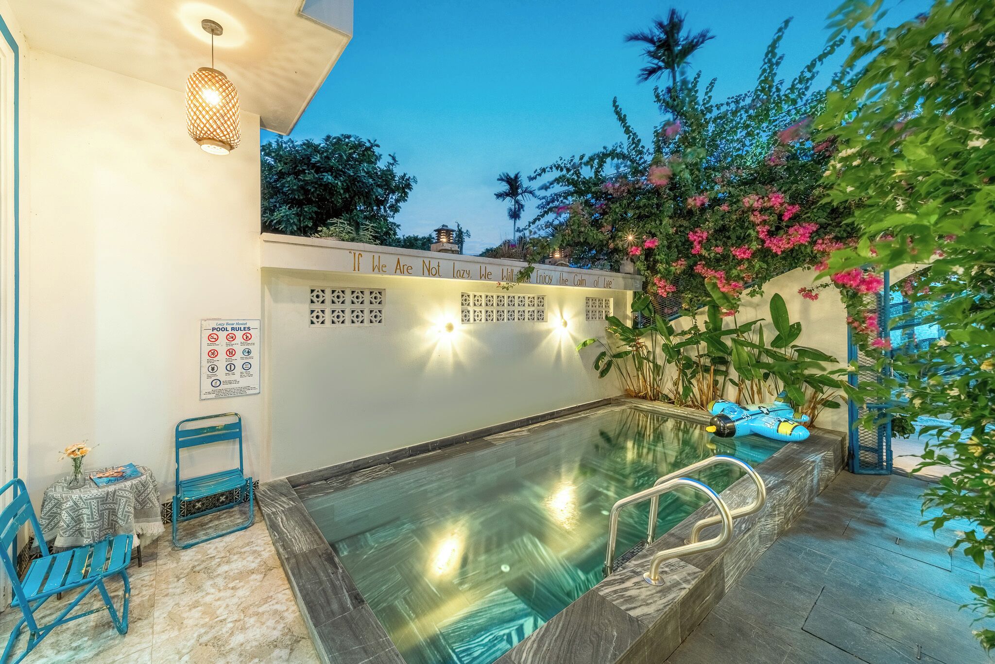 A swimming pool area with a lounge chair, a table with a vase, and a sign with pool rules.
