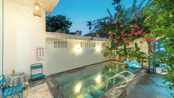 A swimming pool area with a lounge chair, a table with a vase, and a sign with pool rules.