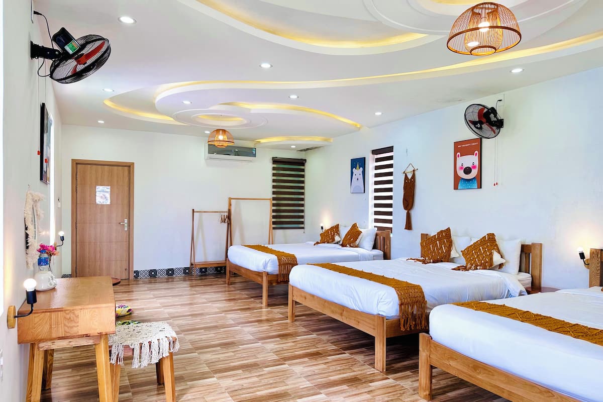 A hotel room with two beds, a wooden desk, a standing fan, a ceiling light fixture, and a wall-mounted air conditioner.