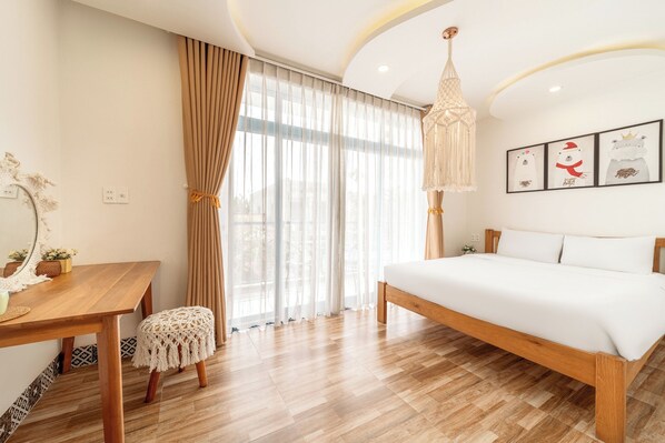 Une chambre à coucher avec un lit en bois, un bureau en bois, un petit tabouret, une grande fenêtre avec des rideaux et un luminaire suspendu.