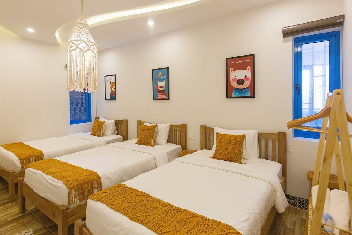 A hotel room with four single beds, a wooden headboard, a hanging macrame light fixture, and a window with a blue frame.