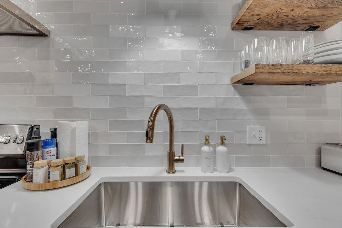 A modern kitchen with a stainless steel sink, a wooden shelf above the sink, and a countertop with jars of spices and a toaster.