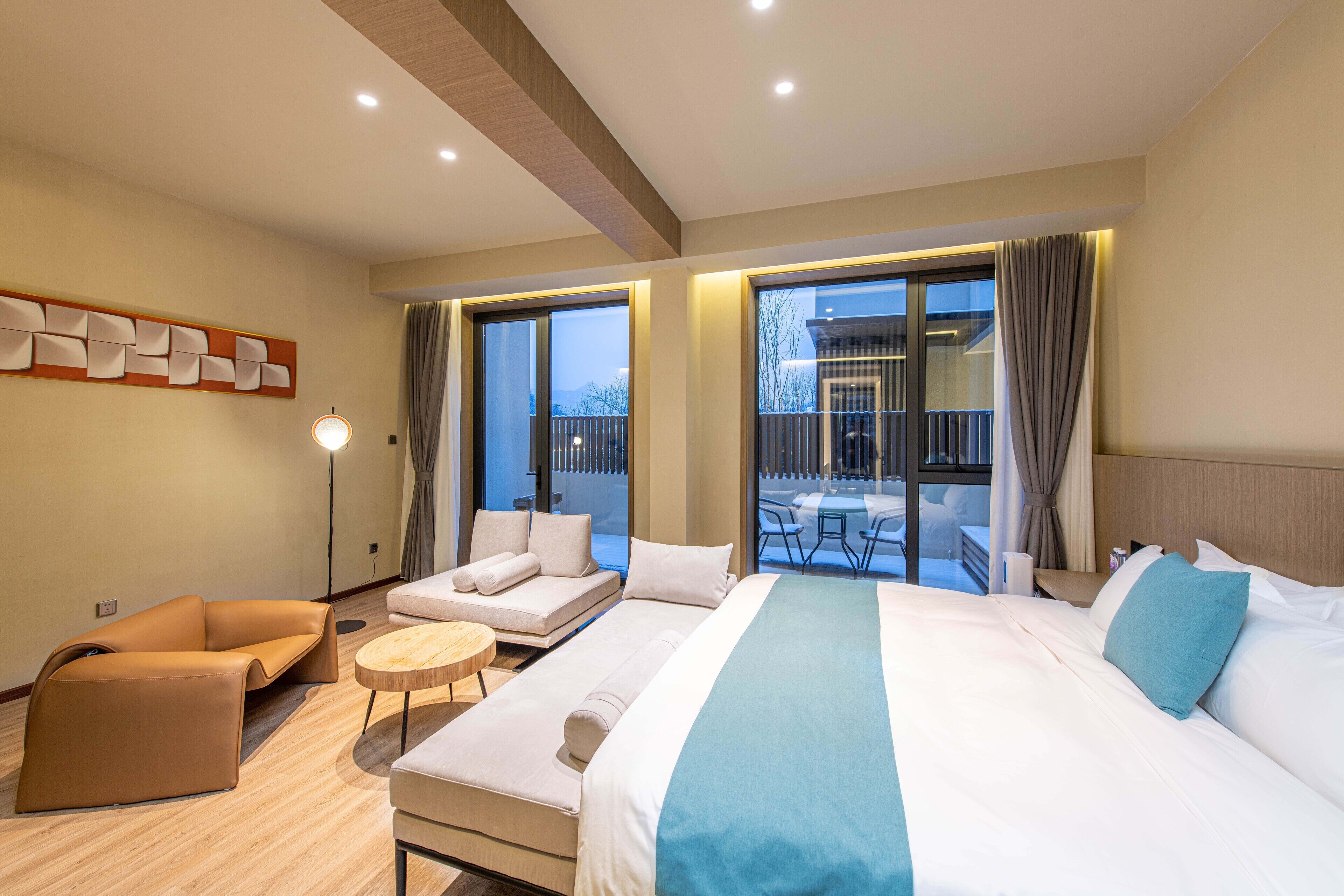 Una habitación de hotel moderna con una cama grande, un sofá, una mesita redonda y un balcón con mobiliario exterior.