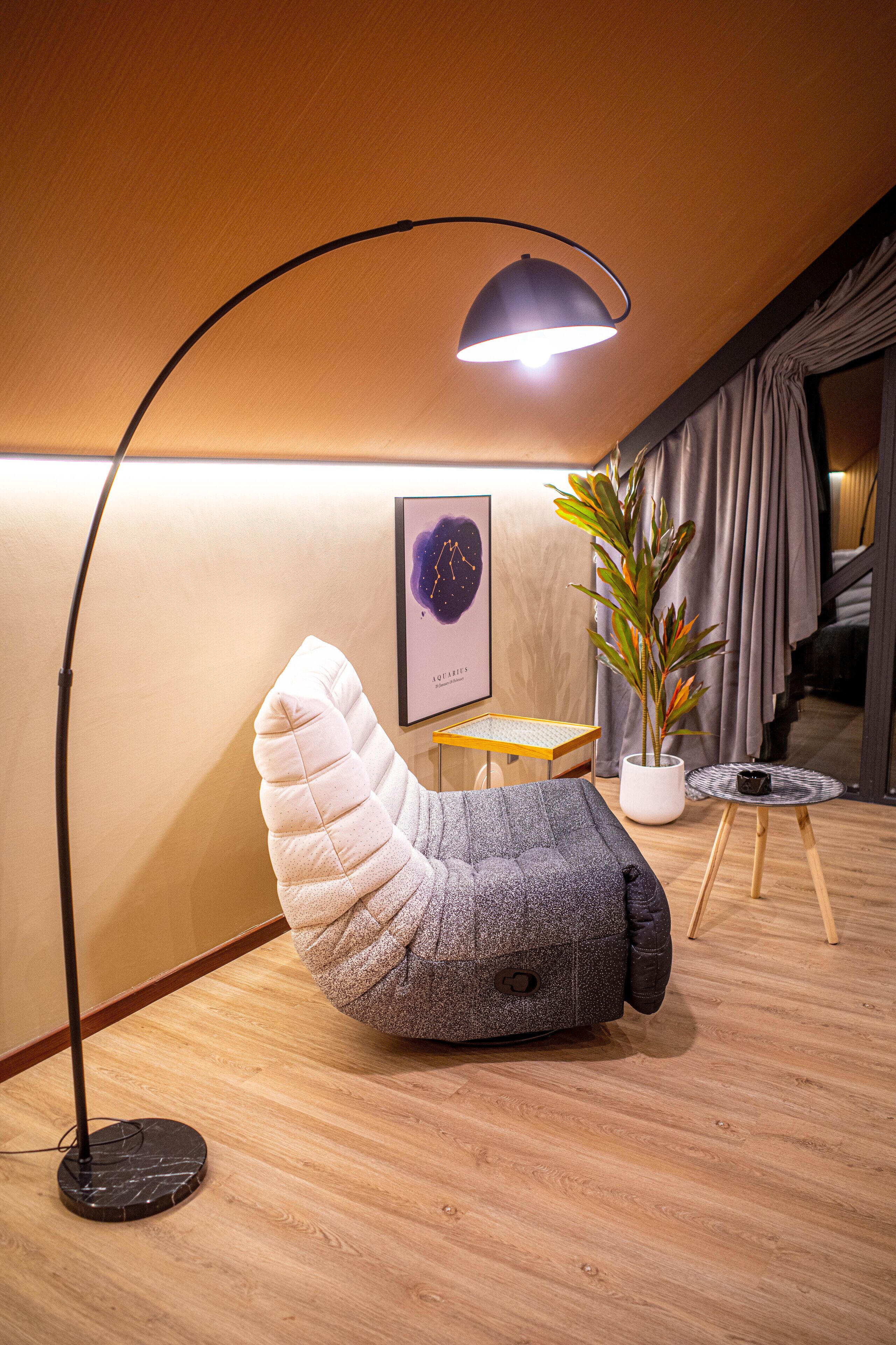 Um quarto aconchegante com poltrona de feltro, luminária de chão, mesa pequena, planta em vaso e quadro na parede.