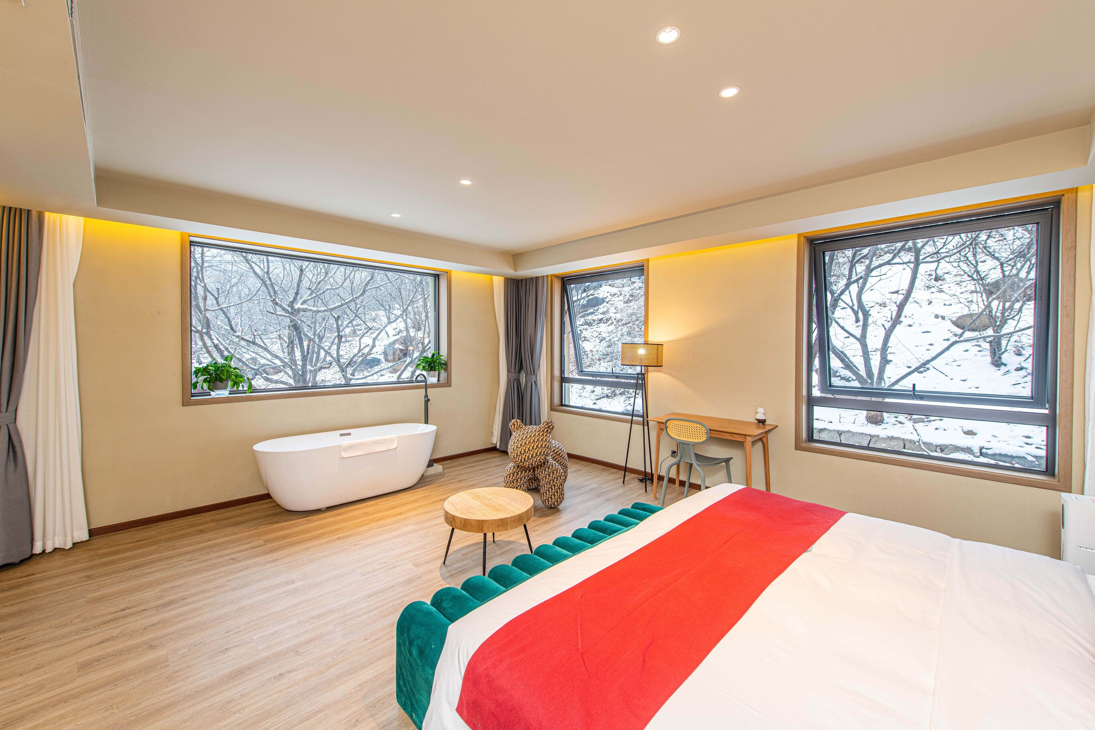 A modern bedroom with a large bed, a bathtub, and two windows with views of trees.