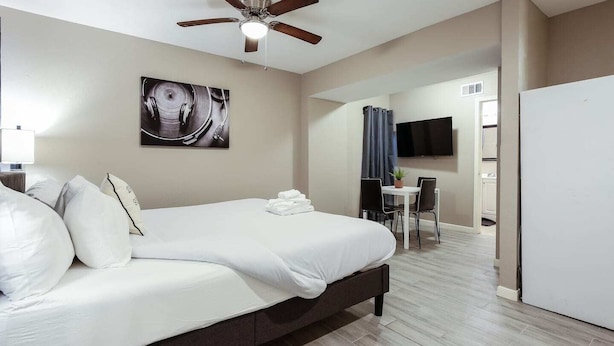 A modern hotel room with a large bed, a ceiling fan, a flat-screen TV, a dining area with a table and chairs, and a kitchen visible through an open door.