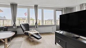 A modern living room with a large window, a black leather chair, a flat-screen TV, and a wooden dining table.