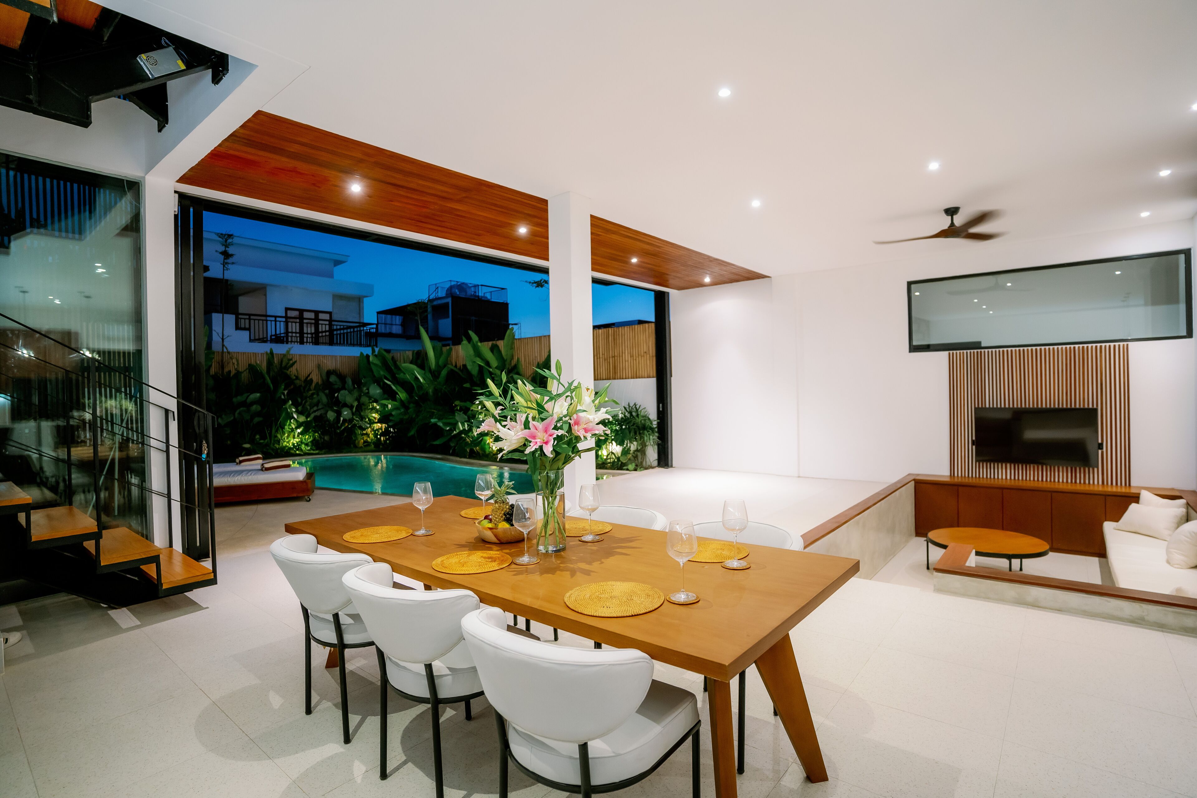 A modern dining area with a wooden table and white chairs, a TV mounted on the wall, and a pool visible through the glass doors.