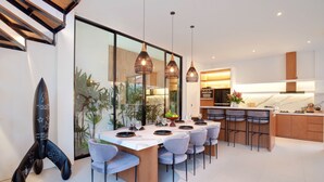 A modern kitchen with a central island, pendant lighting, and a dining area with a long table and chairs. - Paradise Living Villa Canggu (Canggu)