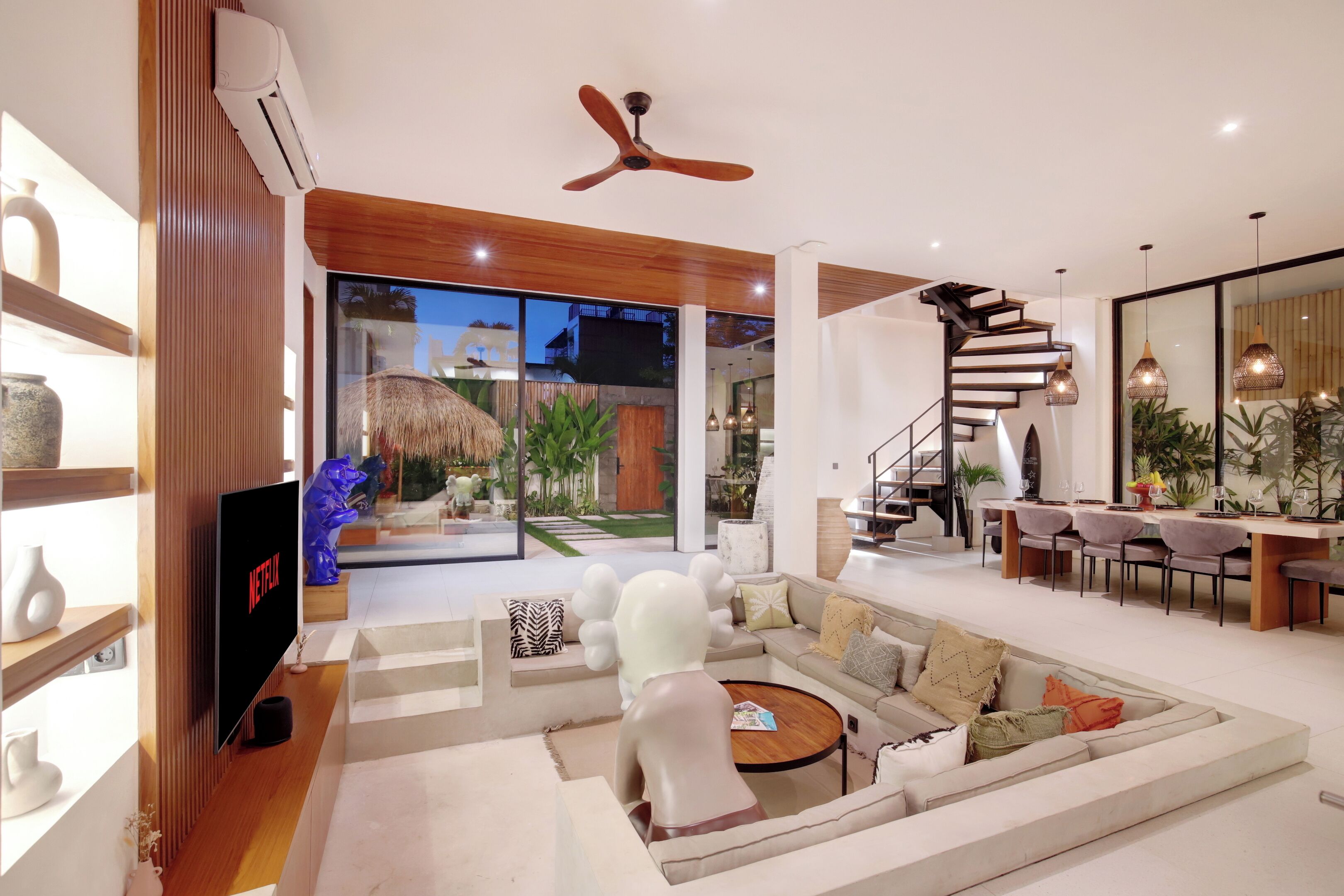 A modern living room with a sectional sofa, a coffee table, and a TV displaying 'Netflix'. There is a staircase leading to another level, and a dining area with chairs and a table.