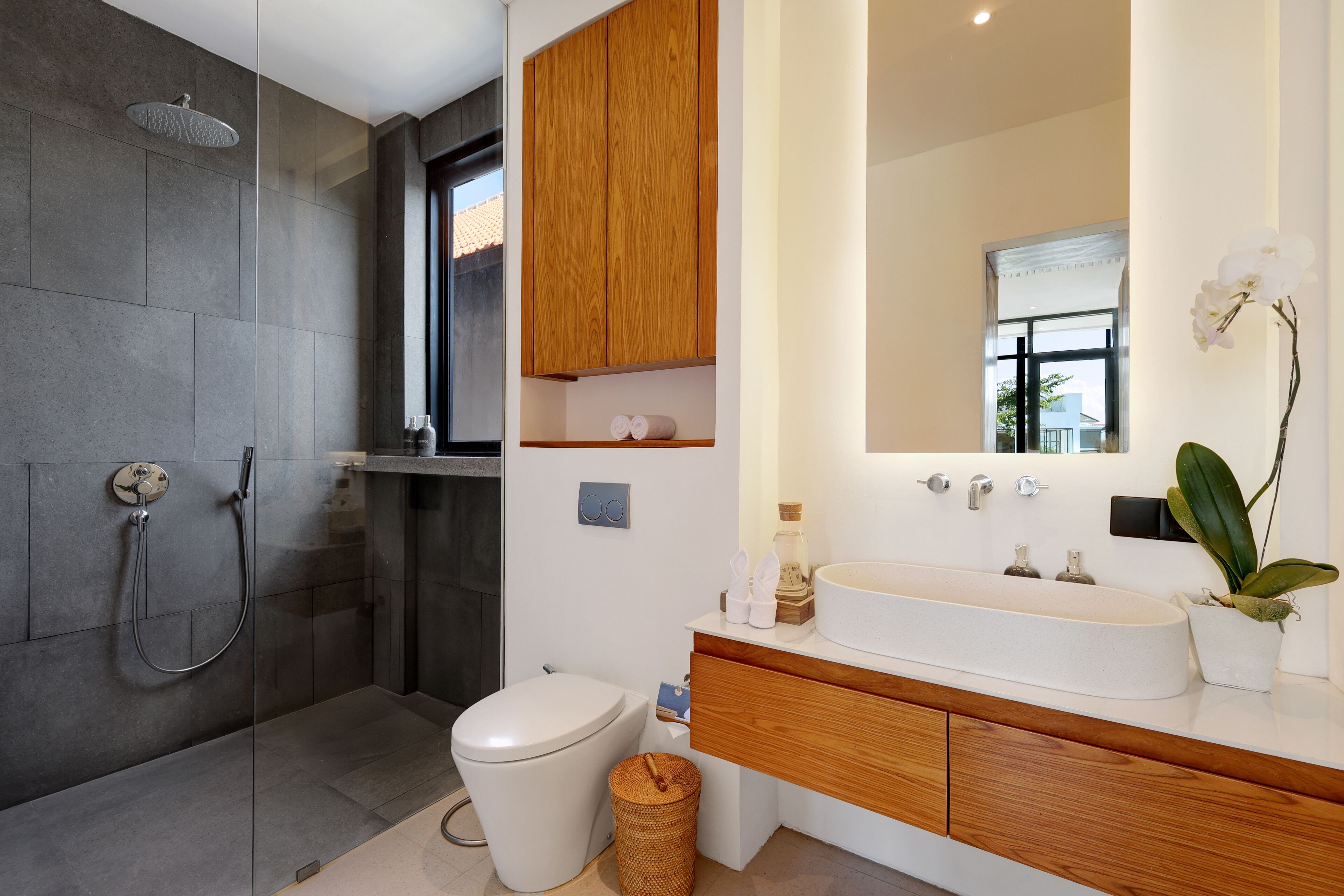 Een moderne badkamer met een douchecabine van glas, een wit toilet, een houten wastafelmeubel met een grote spiegel en een potorkidee.
