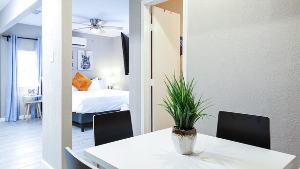 Ein modernes Hotelzimmer mit Bett, Schreibtisch und einem Esstisch mit Stühlen. An der Decke befindet sich ein Ventilator und auf dem Tisch ein Blumentopf.