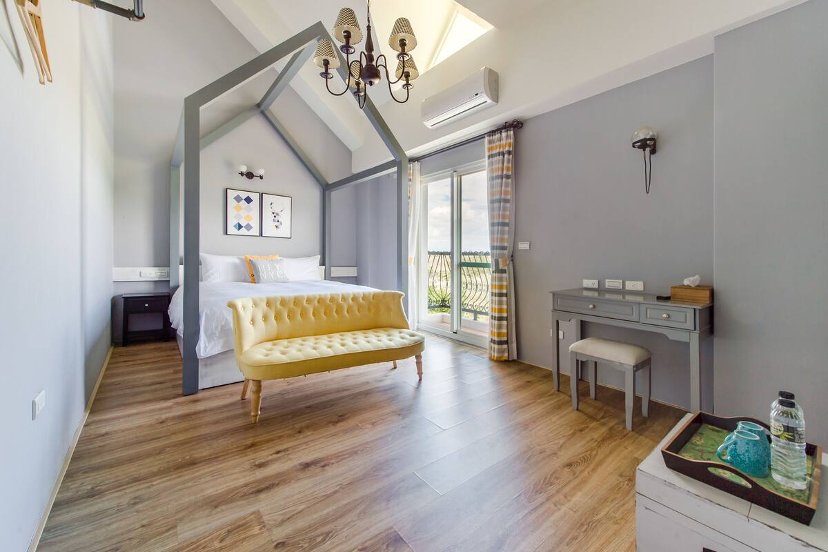A bedroom with a bed, a bench, a desk, and a chair. There is a window with curtains and a view of the outside.