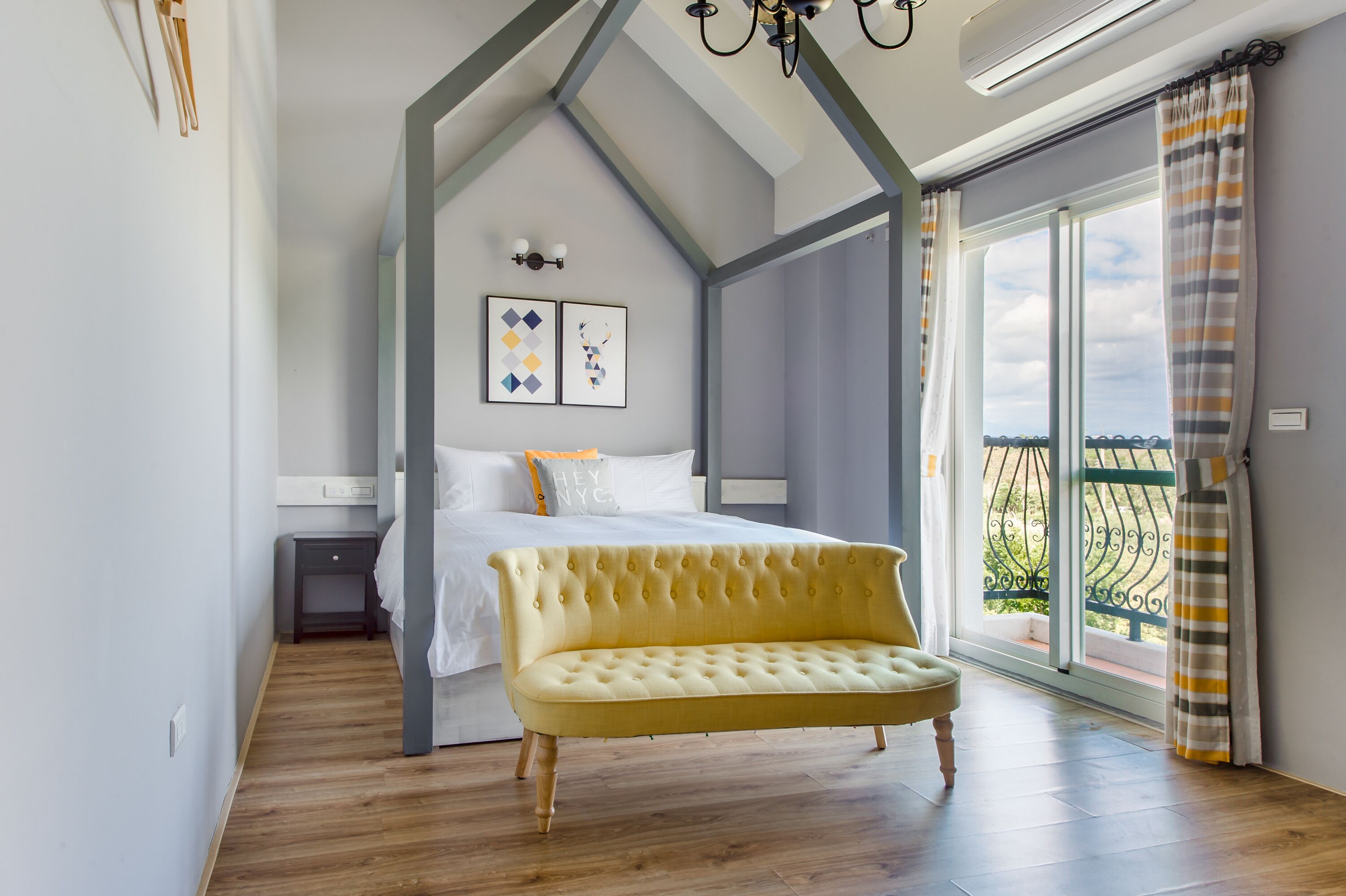 A bedroom with a bed, a yellow bench, a nightstand, and a view of the outdoors through a large window.