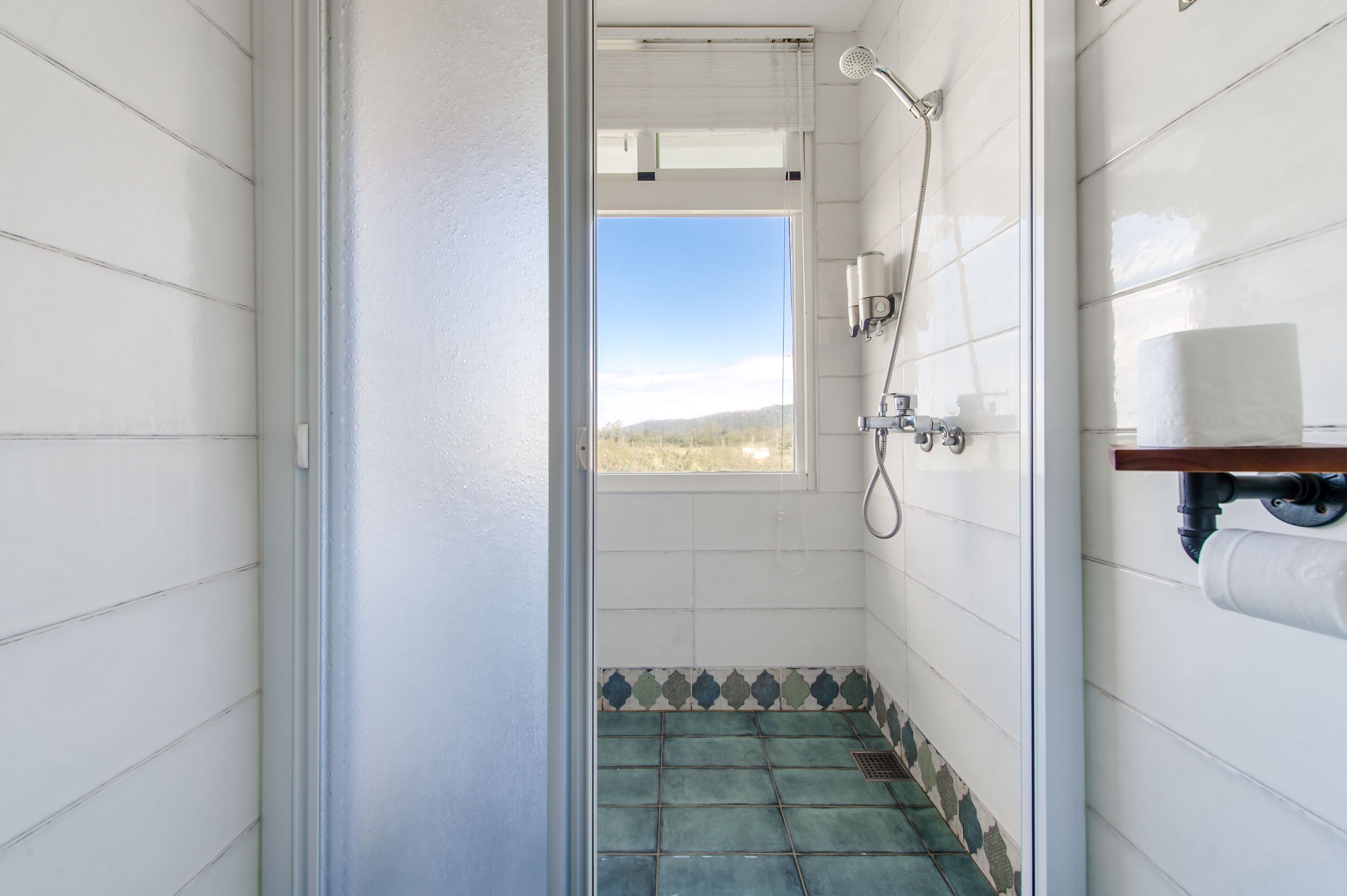 A bathroom with white tiled walls, a shower with a handheld showerhead, a window with a view of a landscape, and a wooden shelf with a roll of toilet paper.