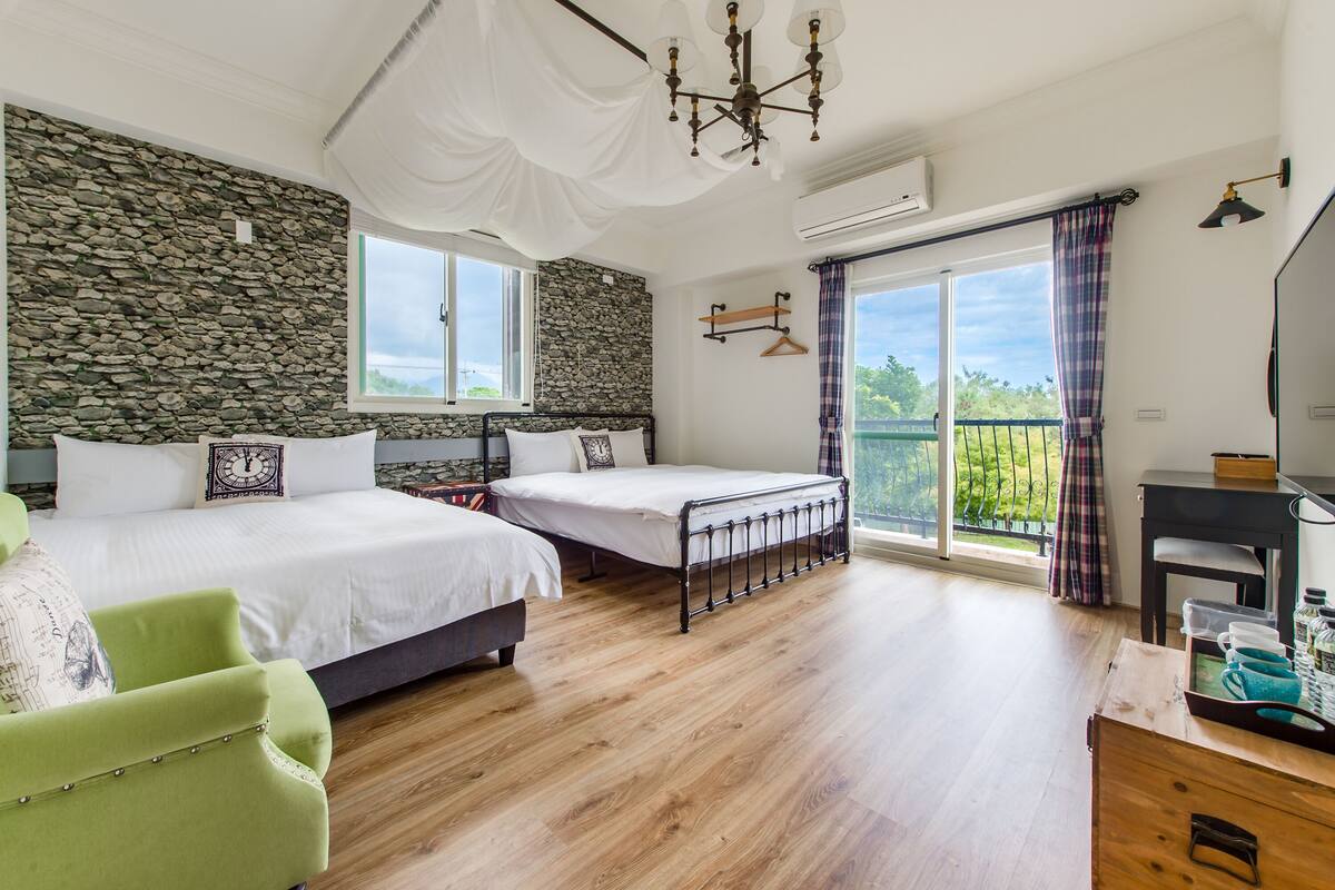 A bedroom with two beds, a green armchair, a desk with a chair, a stone wall, and a balcony with a view.