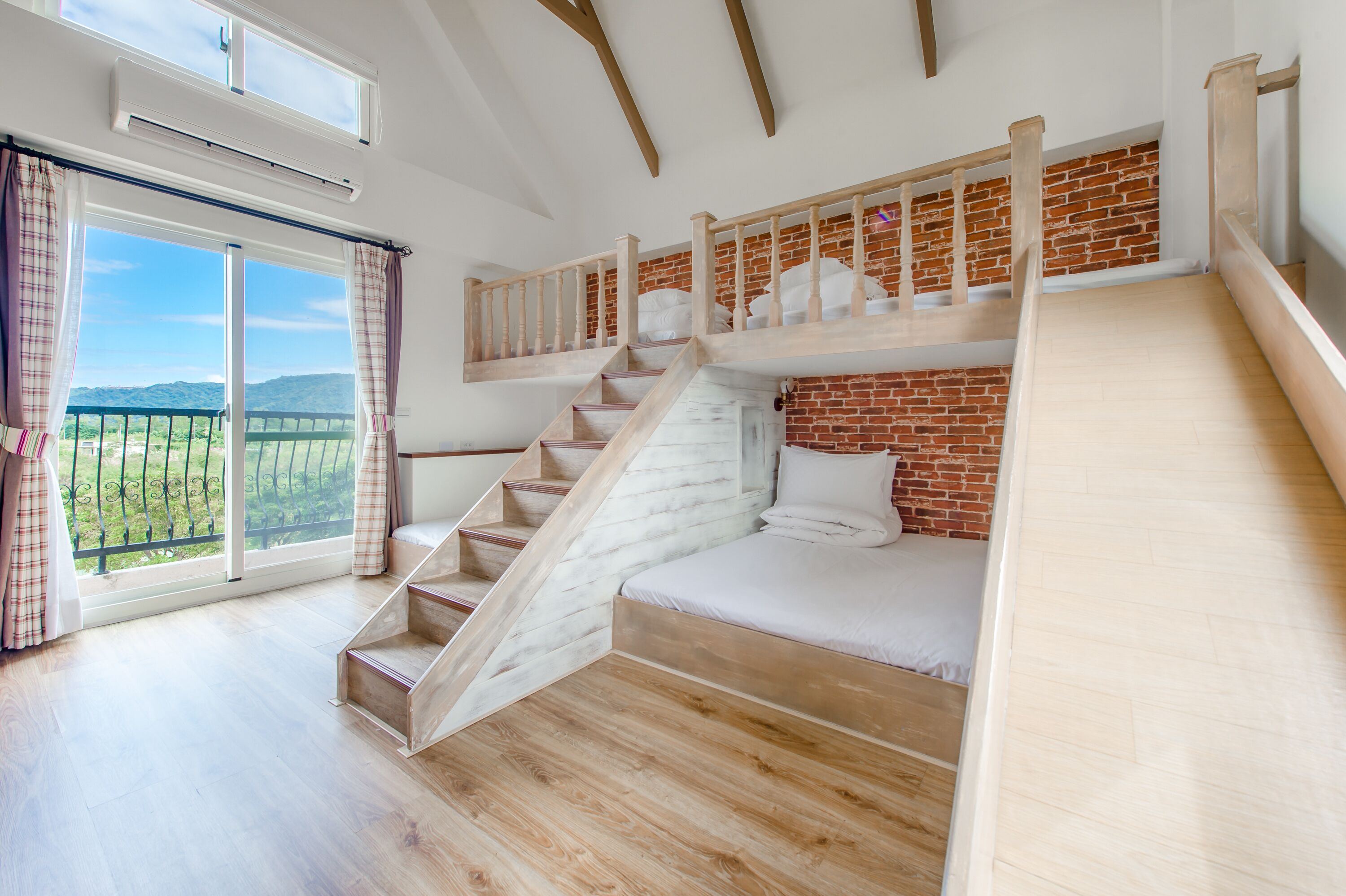 A bedroom with a wooden staircase, a brick wall, and a large window with curtains.