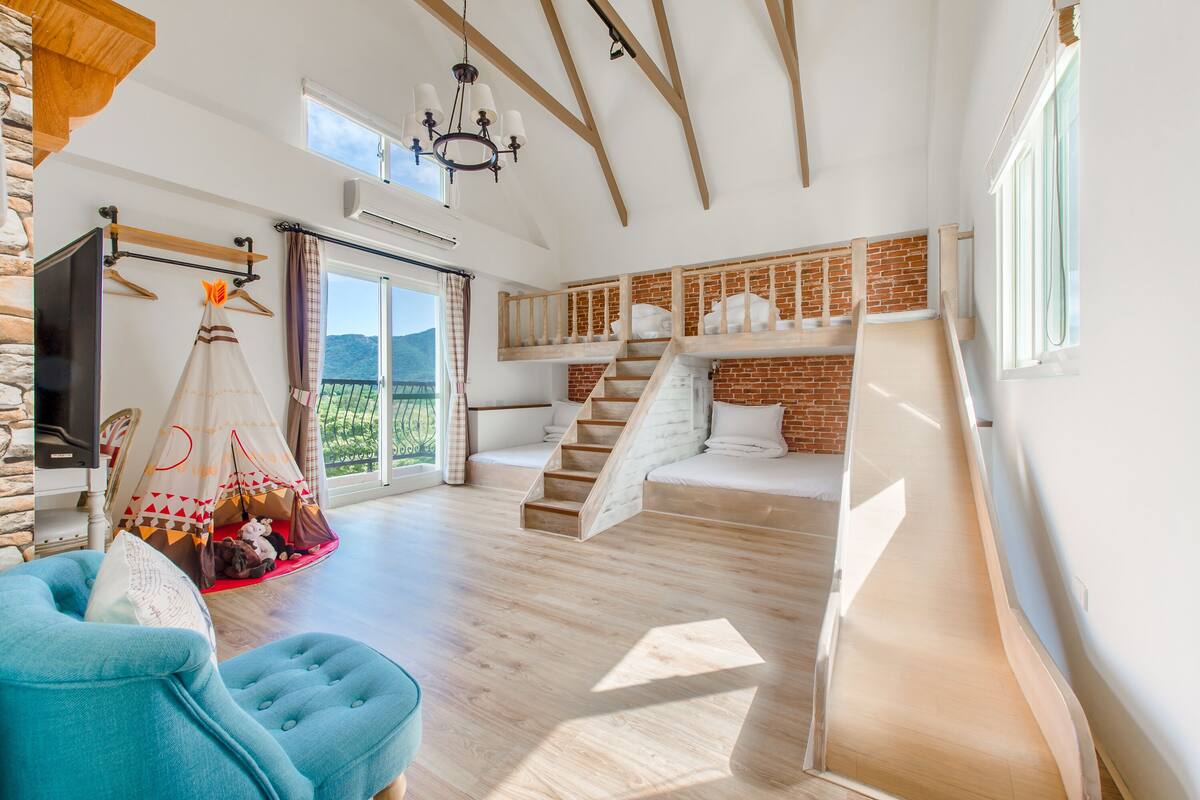 A spacious room with a wooden floor, a blue chair, a white sofa, a tent-like structure, and a staircase leading to a loft area.