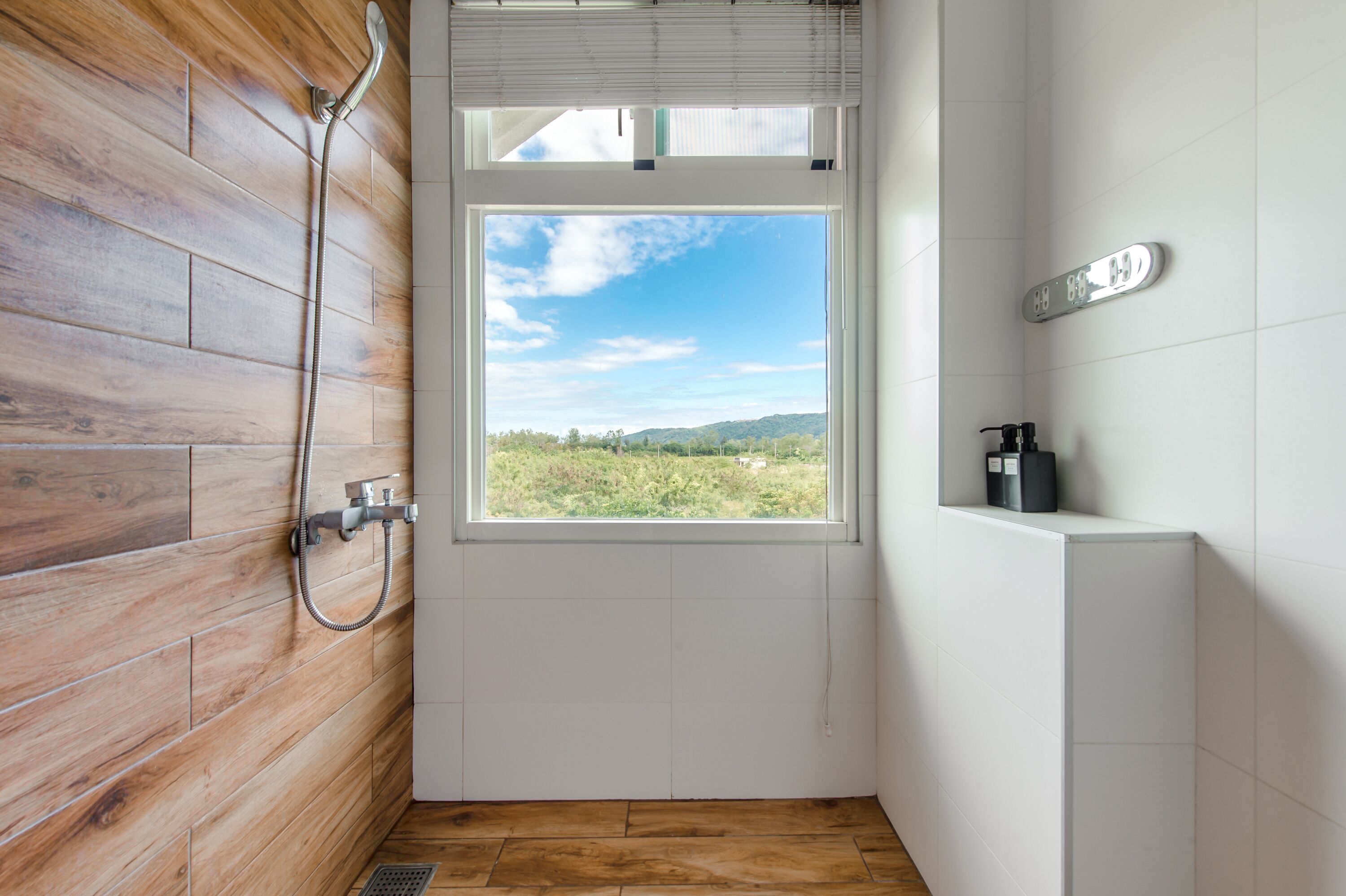 A bathroom with a wooden wall, a shower, a window with a view of greenery, and a white sink.