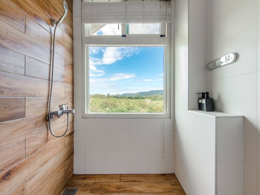 A bathroom with a wooden wall, a shower, a window with a view of greenery, and a white sink.