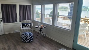 Een kamer met uitzicht op een meer, een stoel met een kussen met de tekst 'MEER', een kleine tafel met een bord en een deur.