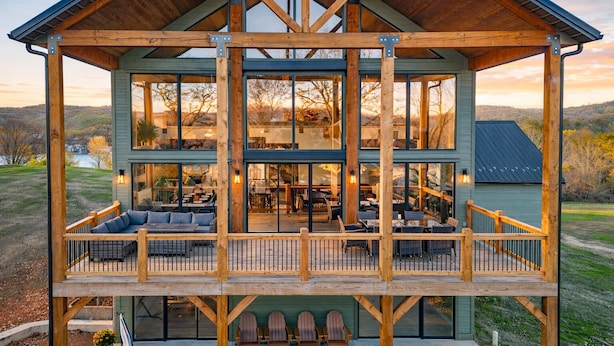 A two-story wooden cabin with a balcony, large windows, and a patio area with seating. - Big Family Lake Retreat | 6BR Table Rock (Cape Fair)