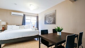 Hotellihuone, jossa on sänky, ruokapöytä tuoleineen, ruukkukasvi, seinälle asennettu ilmastointilaite ja seinällä oleva kehystetty kuva.