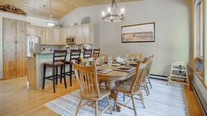 A dining area with a wooden table, chairs, and a chandelier. There is a kitchen in the background with wooden cabinets and a refrigerator.