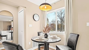 A dining area with a round table, chairs, a clock, and a window with a view of trees. - Charming New Unit in the Heart of Roseville (Roseville)