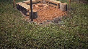 Ein hölzerner Außenbereich mit Sitzgelegenheiten und einem Feuerkorb, umgeben von einer Pergola mit Lichterketten.