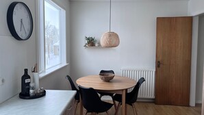 Et spiseområde med et rundt træbord, sorte stole, et vægur, en hængende lampe og et vindue med udsigt til bygninger.