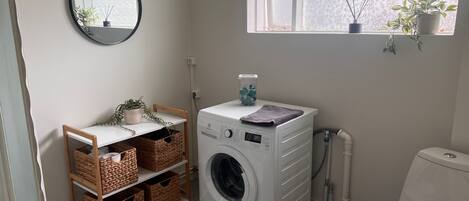 A laundry room with a front-loading washing machine, wicker baskets on shelves, a mirror, and a window with plants.