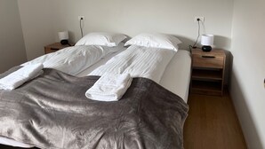 En pænt redt tvillingeseng med hvide sengetøj og et gråt tæppe, to sengeborde med lamper, og en stikkontakt på væggen.