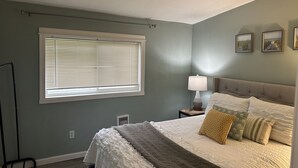 Een slaapkamer met een bed, een nachtkastje met een lamp, een raam met jaloezieën en een wandairconditioner.