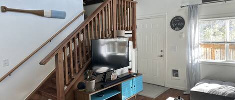 A living room with a wooden staircase, a flat-screen TV, a blue cabinet, and a grey sofa.