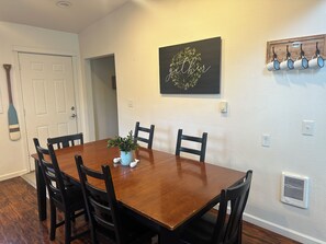 A dining room with a wooden table, six chairs, a potted plant, and a wall-mounted artwork.